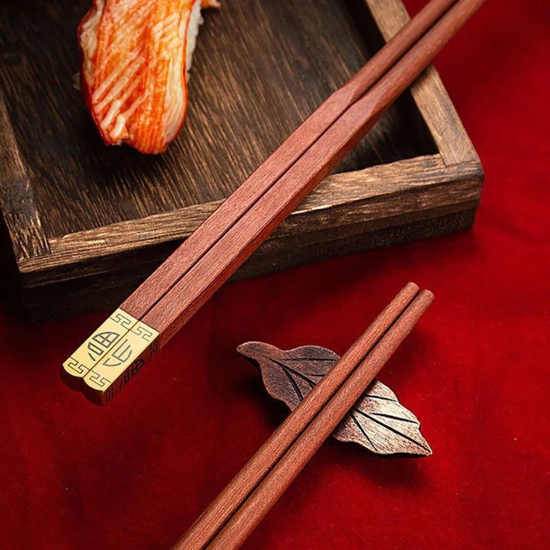 Détail de baguettes chinoises en bois rouge avec motif doré, près d’un repose-baguettes en forme de feuille.